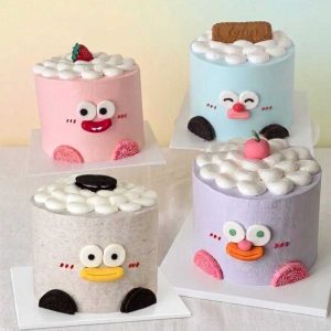 Collection of ugly-cute character cakes in pastel colors featuring playful cartoon faces, fluffy cream dollops, cookies, strawberries, and whimsical decorations by The Cake Alchemy.
